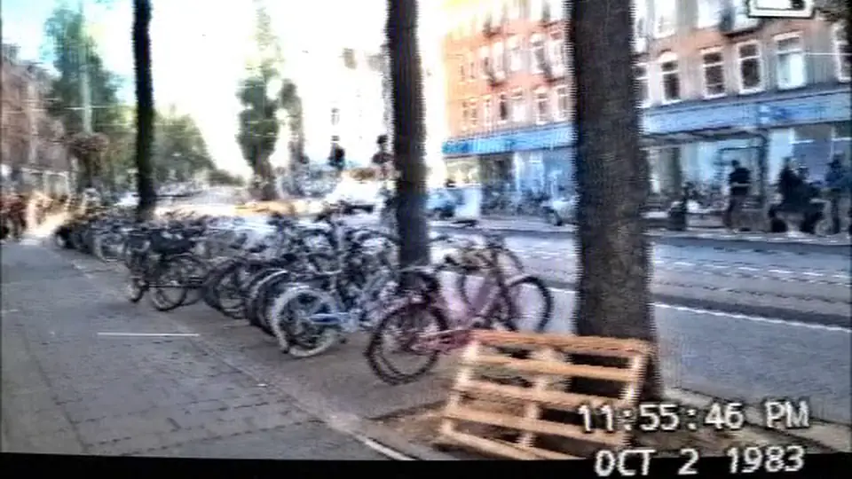 An Amsterdam street with parked bikes, converted to VHS standard play with a 1983 date stamp.